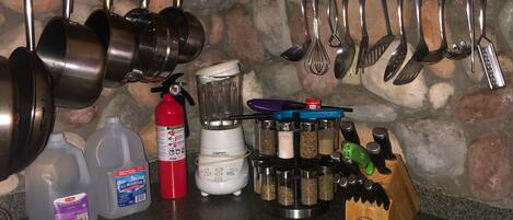 A kitchen with wooden cabinets, a stone backsplash, and various utensils hanging above the counter. There are two bottles of water, a red fire extinguisher, and a blender on the counter.