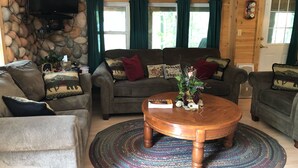 Un salon chaleureux avec un mur en pierre, un plafond en bois et une cheminée. On y trouve deux canapés, une table basse et un téléviseur fixé au mur.