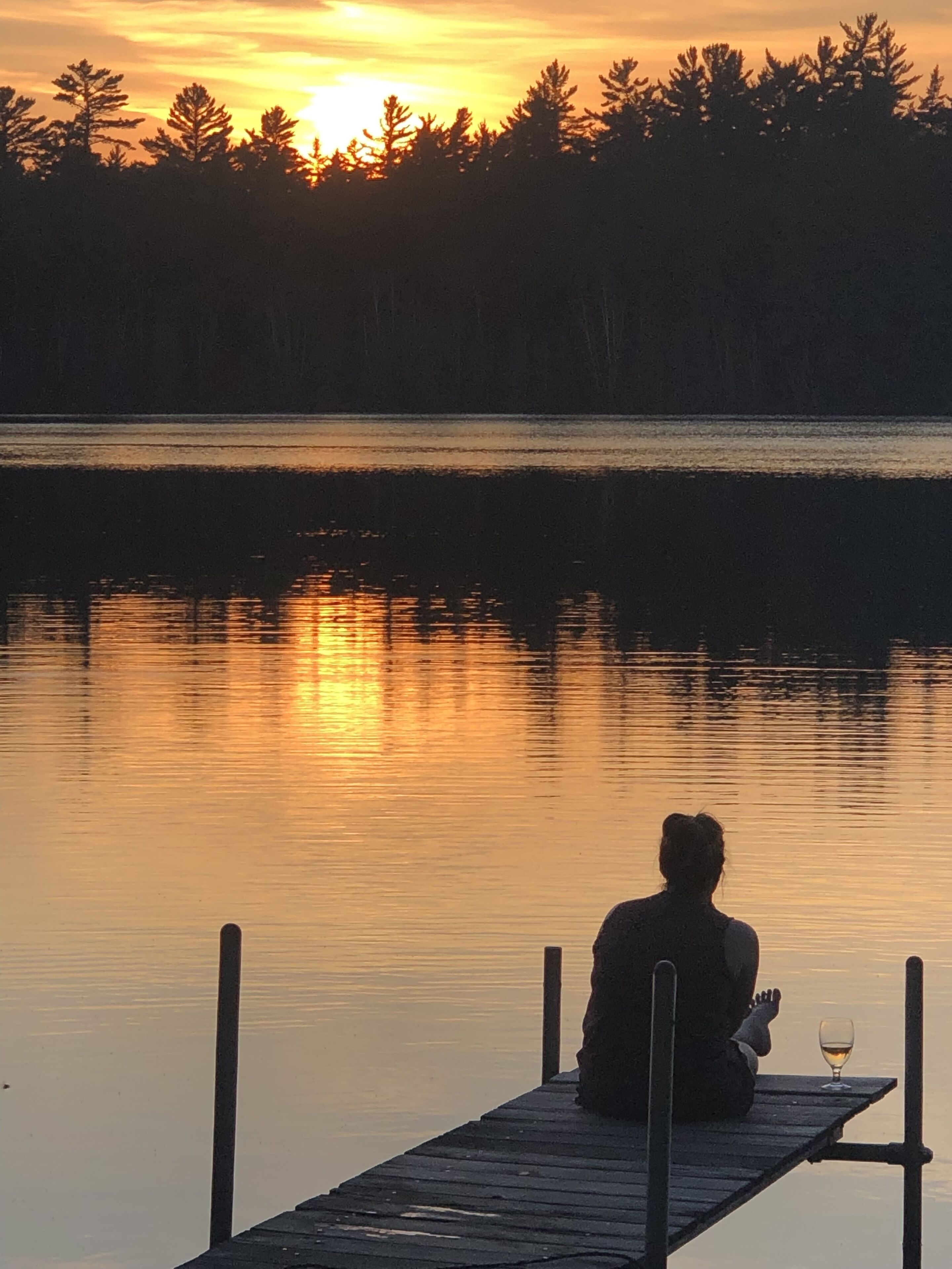 A person sitting on a dock with a glass of wine, overlooking a calm lake with trees in the background.