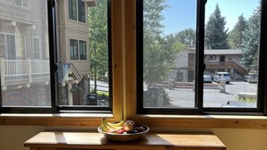 A room with a wooden desk, a bowl of fruit on the windowsill, and a view of a street with buildings and trees.