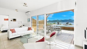 A modern living room with a white sofa, a glass coffee table, and a red chair. There is a large sliding glass door leading to a balcony with a view of a lake and mountains. - Oceanfront amazing 2 BR Villa - 2 min from the beach (Beacon Hill)