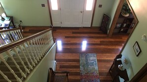 Un couloir doté d’un plancher en bois, d’un escalier avec des balustrades blanches, une porte au fond et une bibliothèque contenant des livres et une plante.