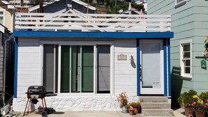 A small, single-story house with a blue front and white shutters, a small porch with a grill, and a stone foundation.