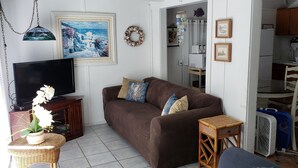 A living room with a brown sofa, a television, a wicker table with a vase of flowers, and a kitchen visible in the background.