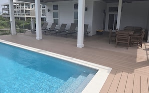 Zona junto a la piscina con sillones de descanso y una mesa, rodeada por una terraza de madera y paredes blancas.