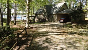 A cabin with a gravel driveway leading to a covered parking area, surrounded by trees and a lake.