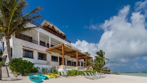 Una proprietà fronte spiaggia con una grande casa, un portico coperto e una spiaggia sabbiosa con lettini.