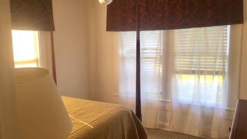 Ein Schlafzimmer mit Bett, Deckenventilator, Fenster mit Jalousien und Blick ins Freie.