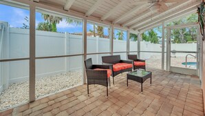 Un patio exterior con mobiliario de mimbre y una mesa de vidrio, rodeado por una cerca blanca y un ventilador de techo.
