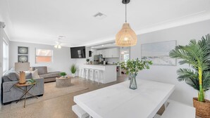 Una sala de estar moderna con un sofá gris, una mesa de comedor blanca y una zona de cocina con armarios blancos y una barra.