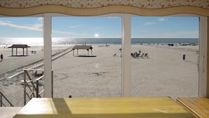 Vista da praia a partir de um quarto, com uma grande janela, mostrando uma praia arenosa, o sol e algumas pessoas.