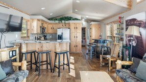 A modern kitchen with wooden cabinets, a central island with stools, and a large TV.