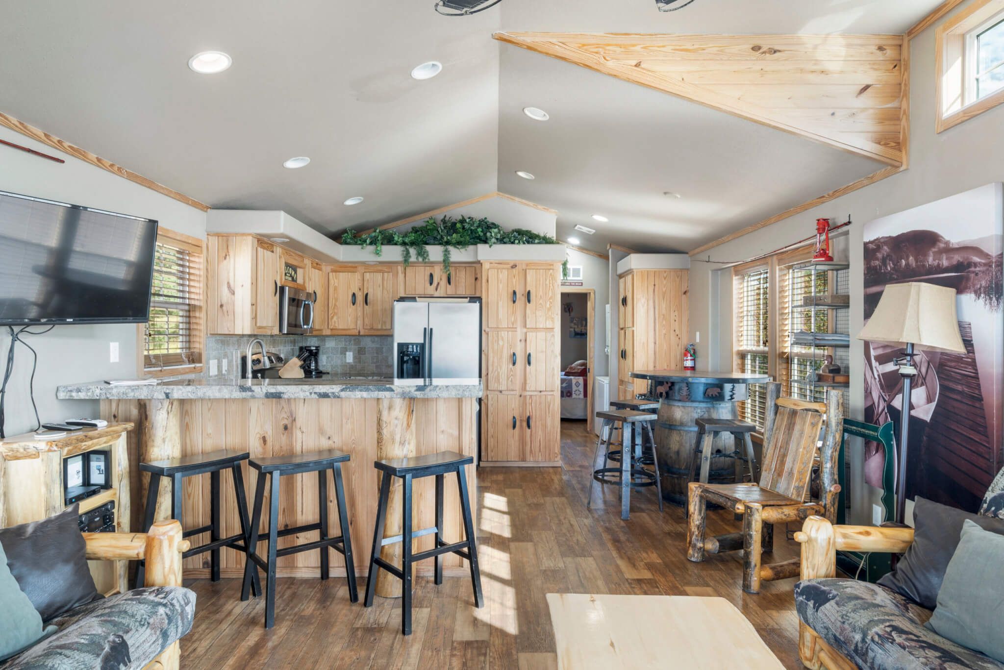 A modern kitchen with wooden cabinets, a central island with stools, and a large TV.