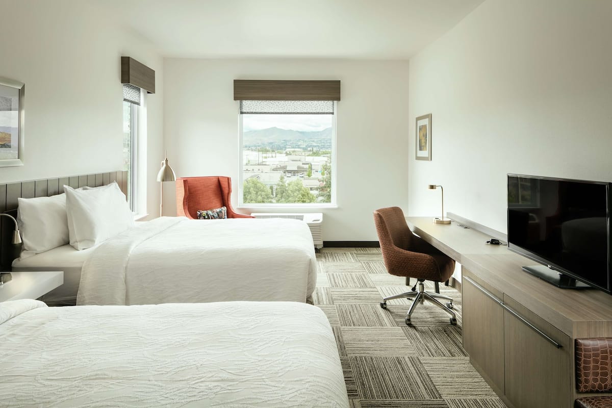 A hotel room with two beds, a desk with a TV, a chair, and a window with a view of buildings and mountains.