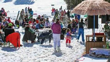 A snowy outdoor gathering with people seated at tables, some under a thatched shelter.