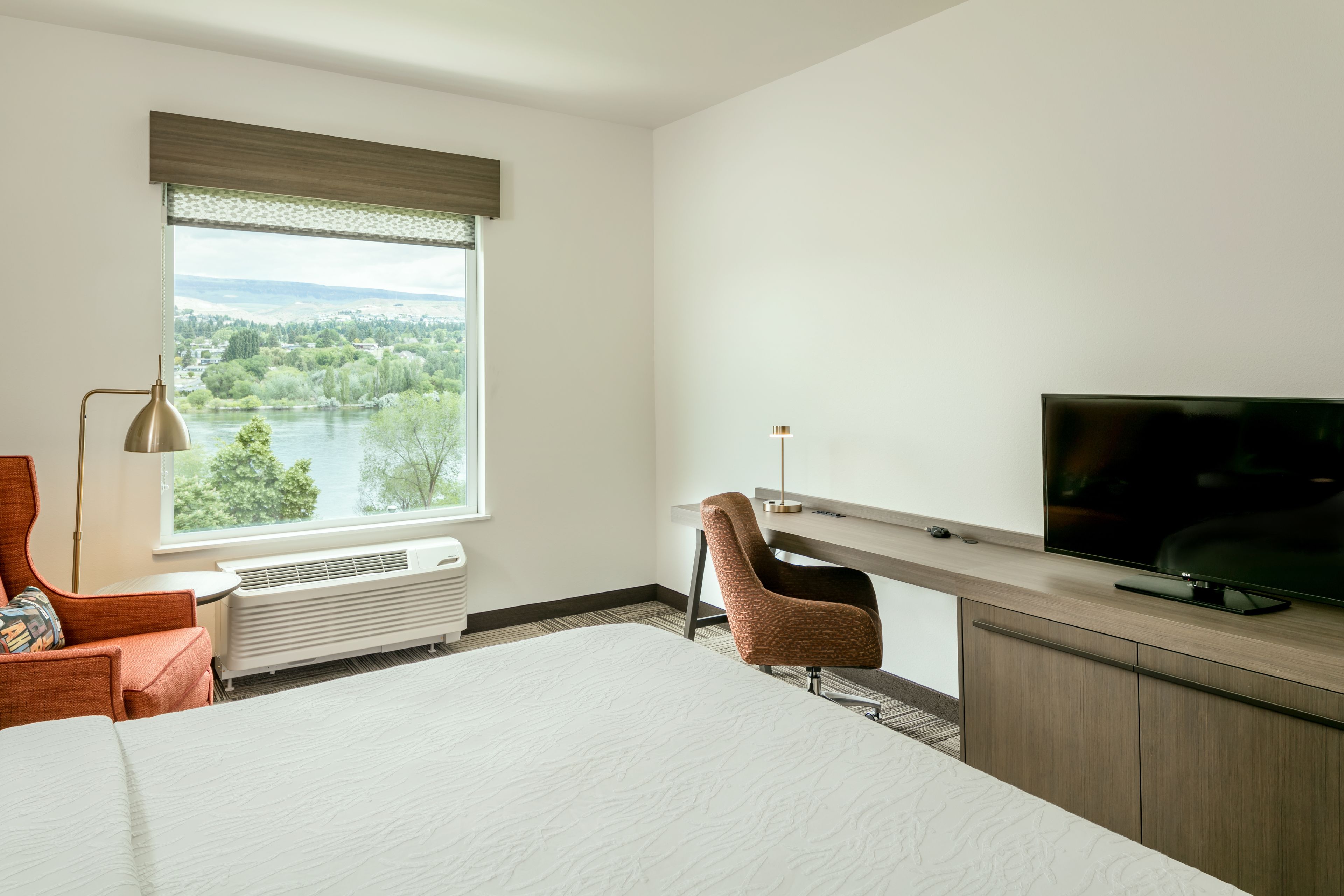 A hotel room with a large bed, a desk with a TV, a chair, a window with a view of trees and water, and a heater.
