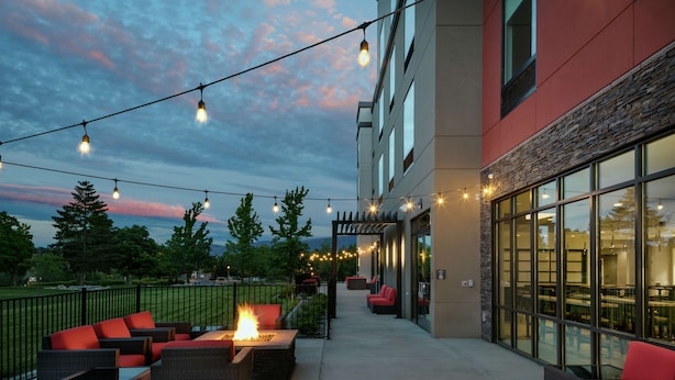 Un espace extérieur aménagé en terrasse, avec des meubles en osier, un foyer et des guirlandes lumineuses, le tout sur fond d’un bâtiment doté de grandes fenêtres et d’une façade en pierre.
