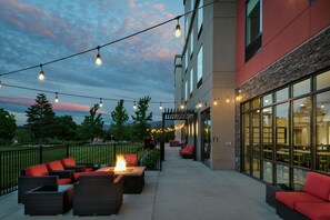An outdoor patio area with wicker furniture, a fire feature, and string lights against a backdrop of a building with large windows and a stone facade. - Hilton Garden Inn Wenatchee (Wenatchee)