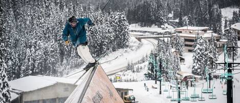 A ski slope with a skier performing a jump, surrounded by snow-covered trees and buildings.