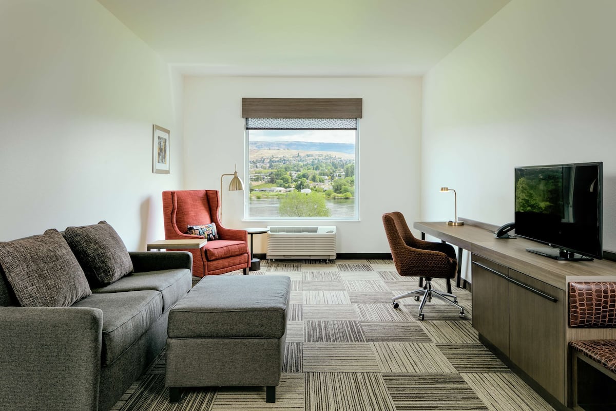 A modern hotel room with a sofa, armchair, and desk with a television.