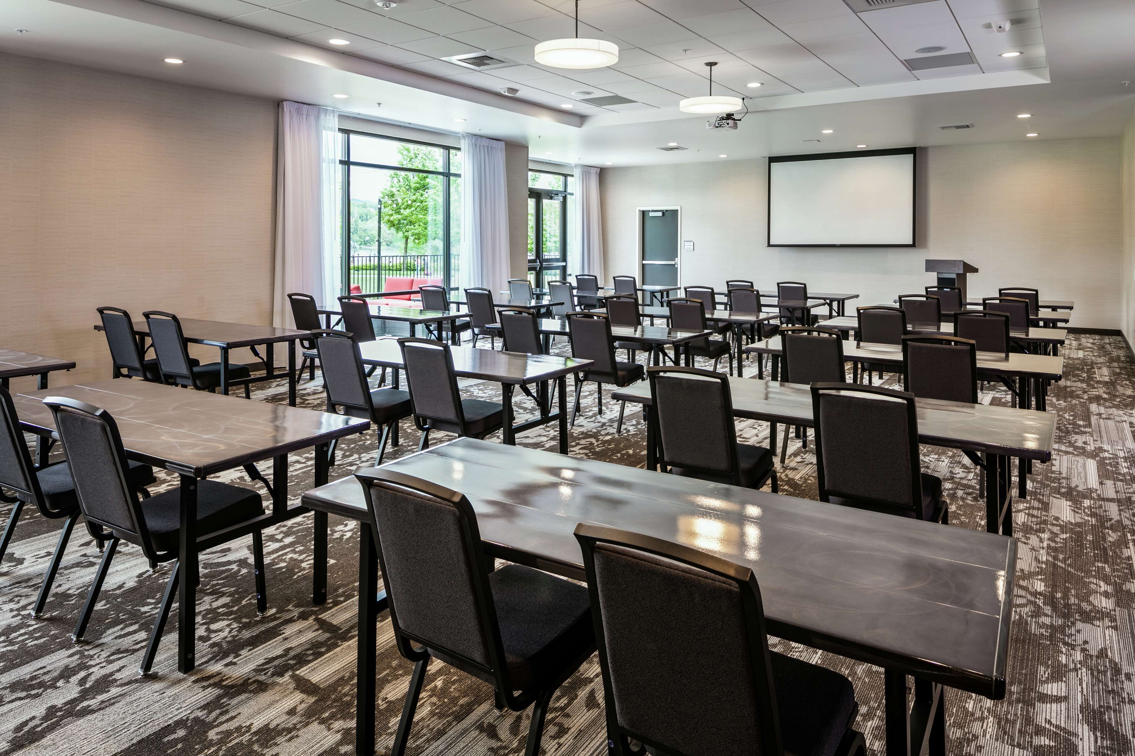 A conference room with multiple tables and chairs, large windows, and a projector screen.