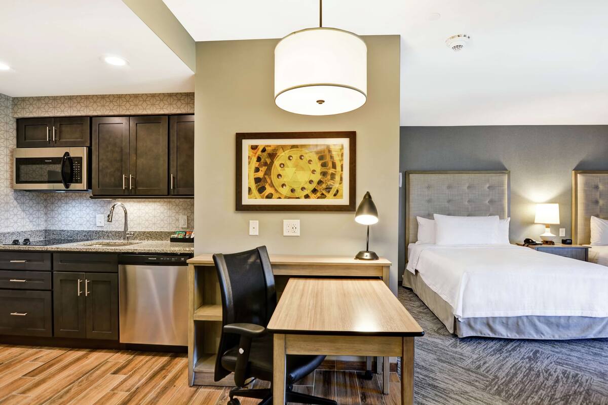 A hotel room with a kitchenette, a dining table with a chair, a bed, and a painting on the wall.