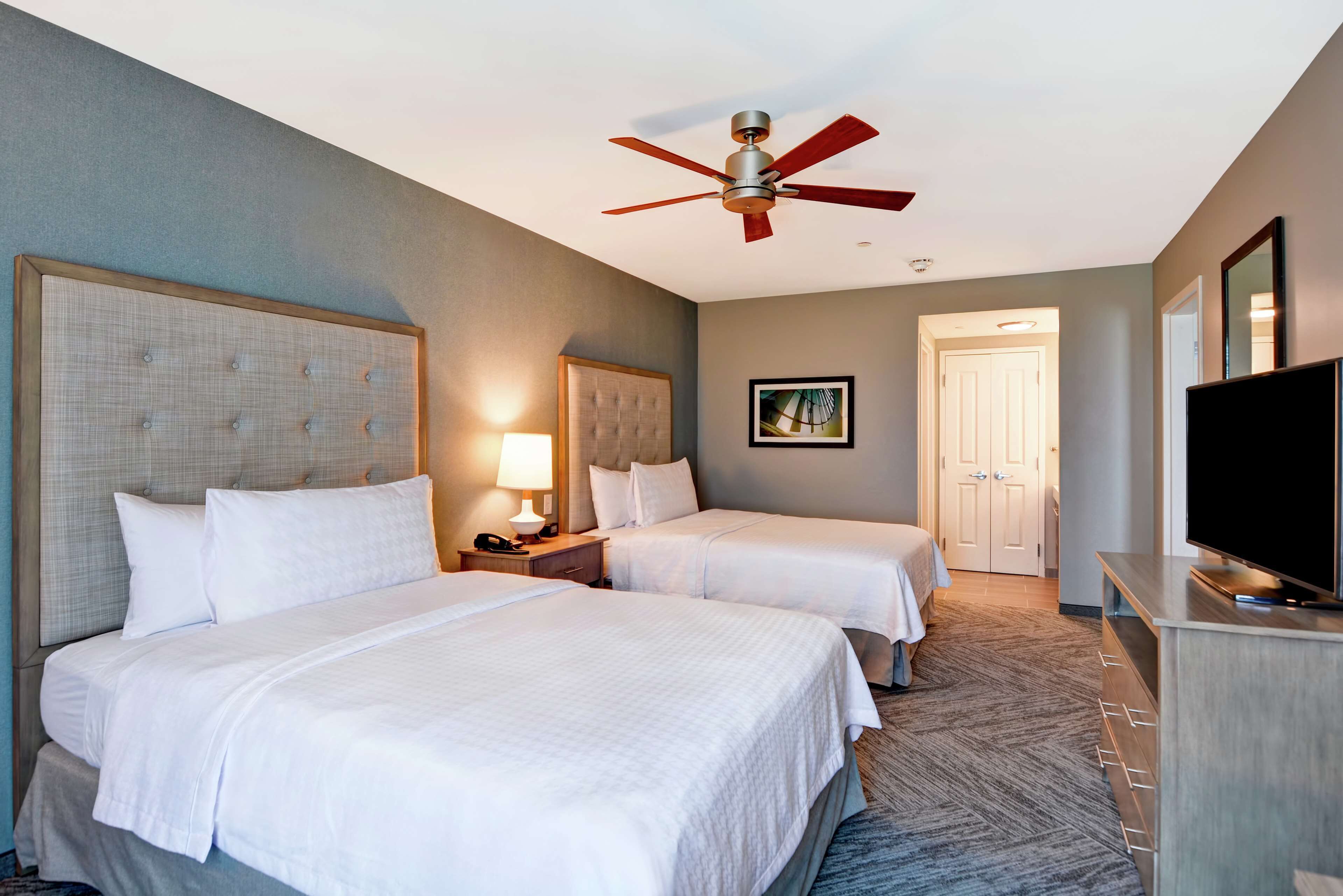 A hotel room with two beds, a ceiling fan, a TV, and a door leading to another room.