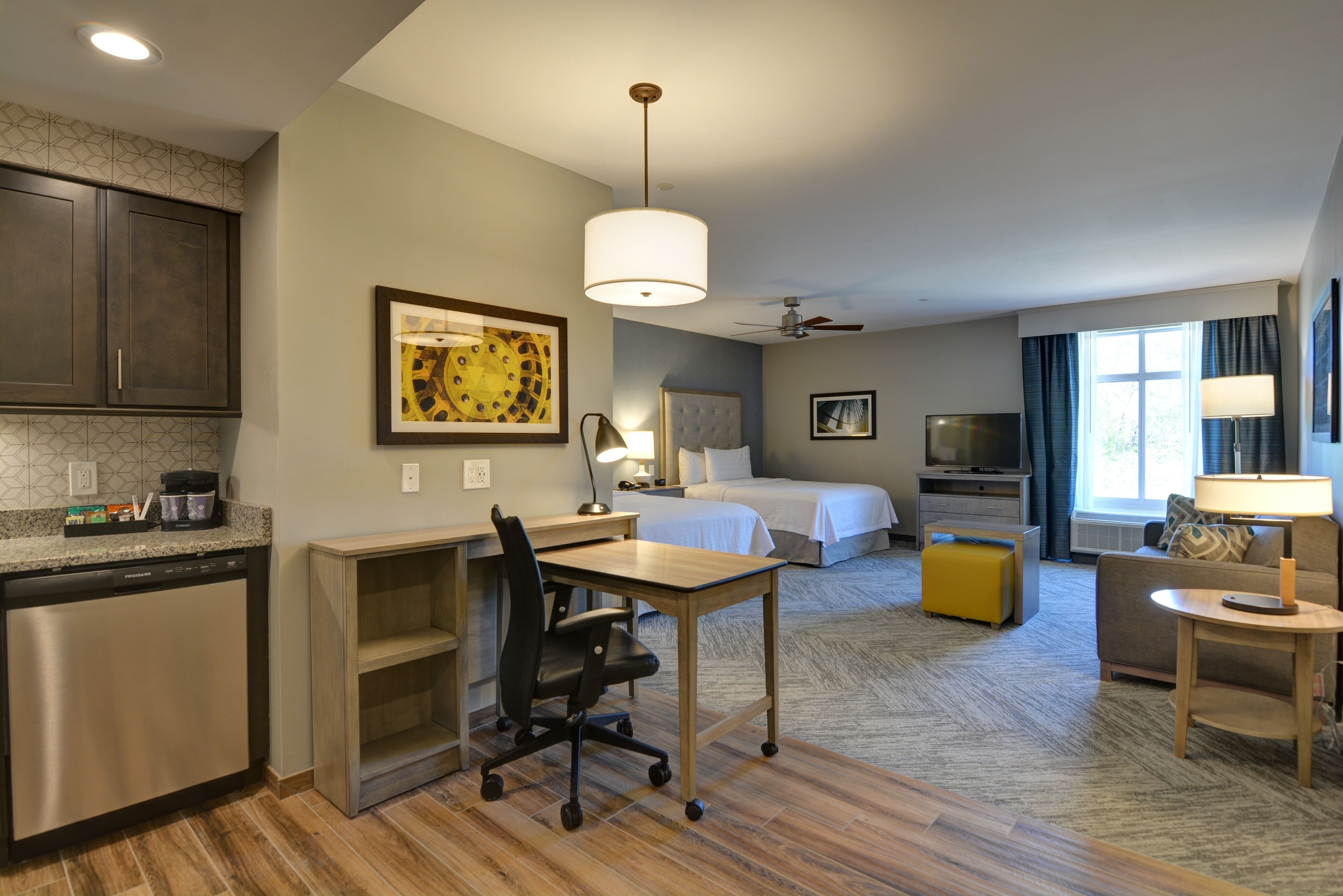 Una habitación de hotel con kitchenette, un escritorio y una silla, una cama, un televisor, un sofá y un ventanal con cortinas.