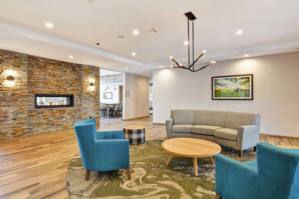 A modern lounge area with a stone wall, a fireplace, a sofa, and two blue armchairs. - Homewood Suites by Hilton Hadley Amherst (Hadley)