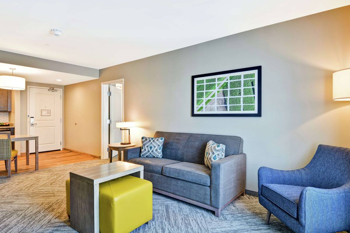 A modern living room with a gray sofa, a yellow ottoman, and a blue armchair. There is a framed artwork on the wall and a lamp on a side table.