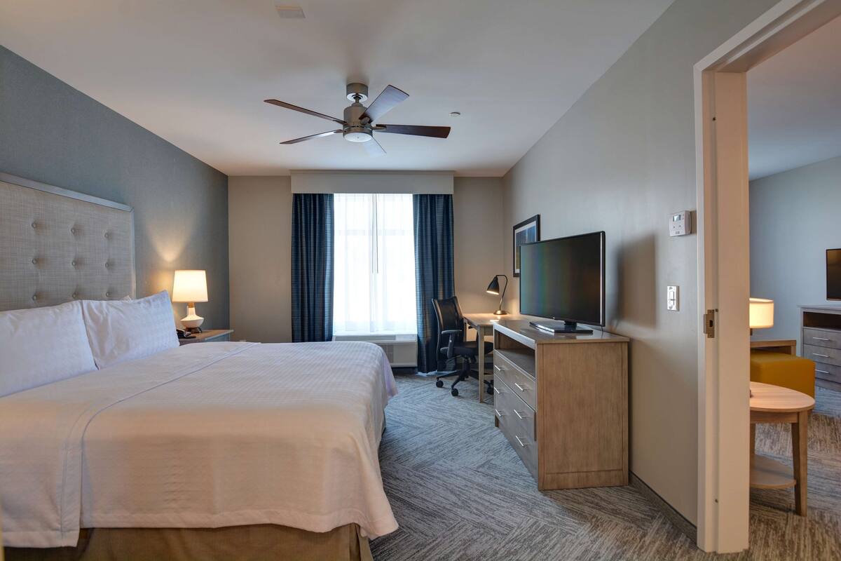 A hotel room with a large bed, a desk with a television, a chair, and a ceiling fan.