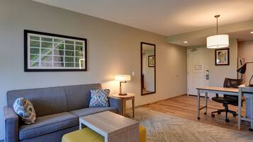 A modern living room with a grey sofa, a yellow ottoman, a small table, and a desk with a chair. There is a framed picture on the wall, a lamp on a side table, and a mirror on the wall.