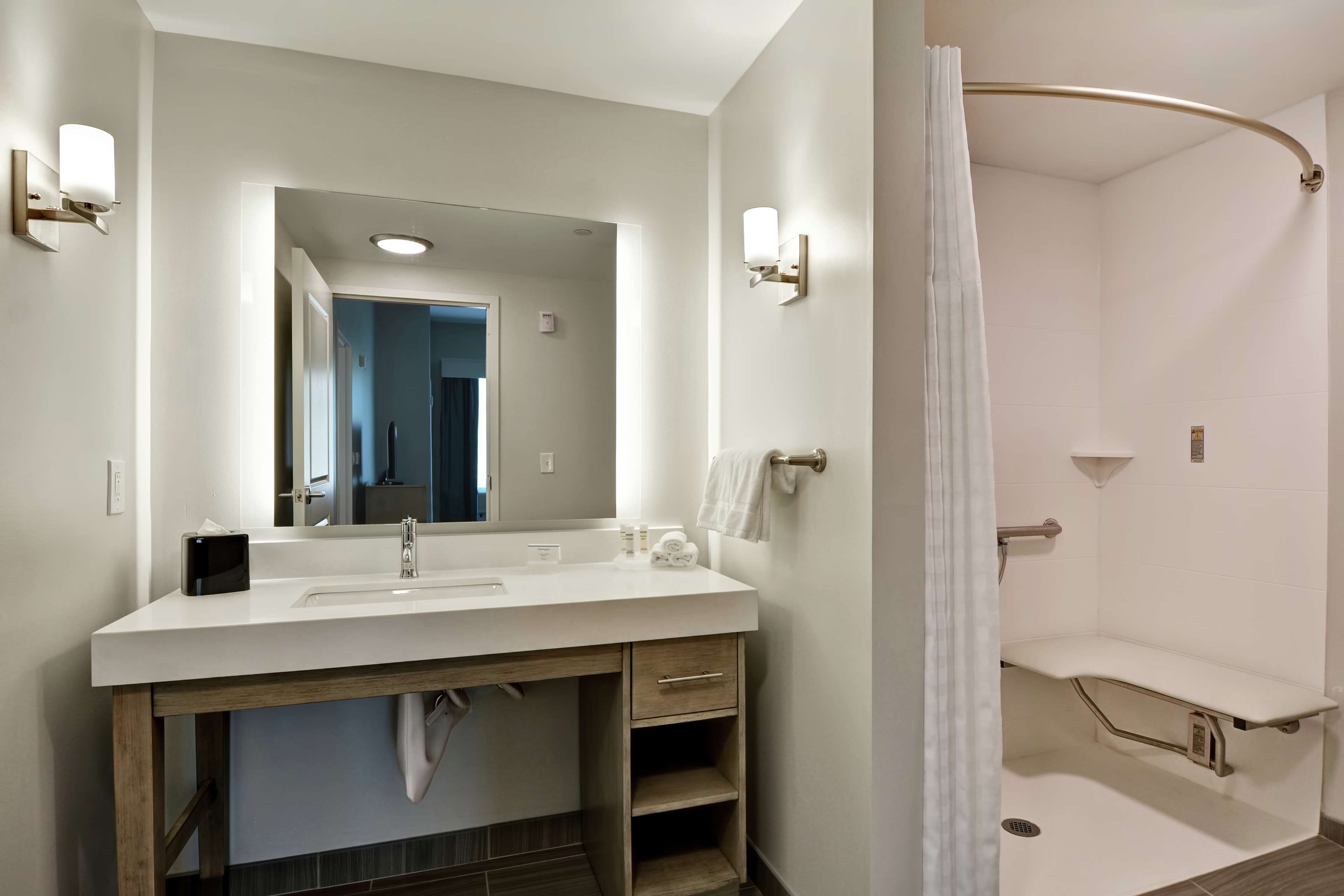 Un baño moderno con un espejo grande, un lavamanos blanco, un perchero y una ducha con puerta de vidrio curvada.