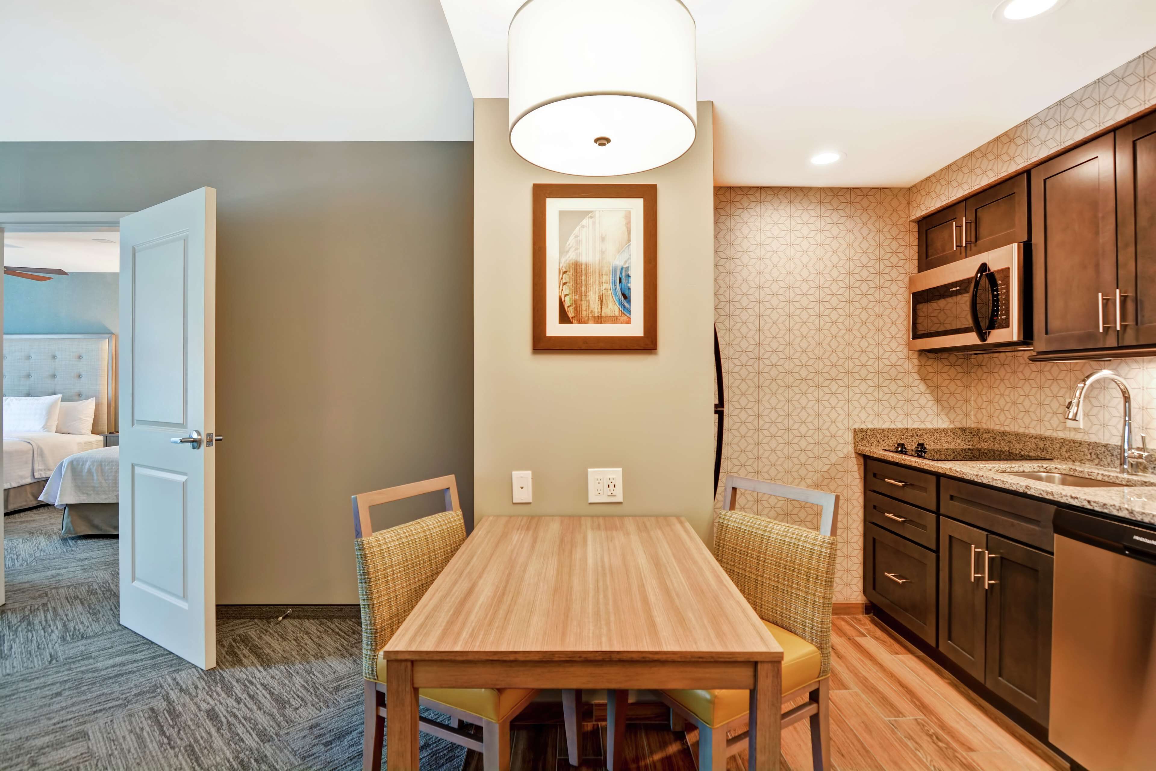Habitación de hotel con comedor que incluye una mesa y sillas de madera, cocina con armarios de madera oscura y electrodomésticos de acero inoxidable, y pared de color neutro con una obra de arte enmarcada.