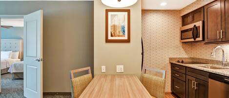 Habitación de hotel con comedor que incluye una mesa y sillas de madera, cocina con armarios de madera oscura y electrodomésticos de acero inoxidable, y pared de color neutro con una obra de arte enmarcada.
