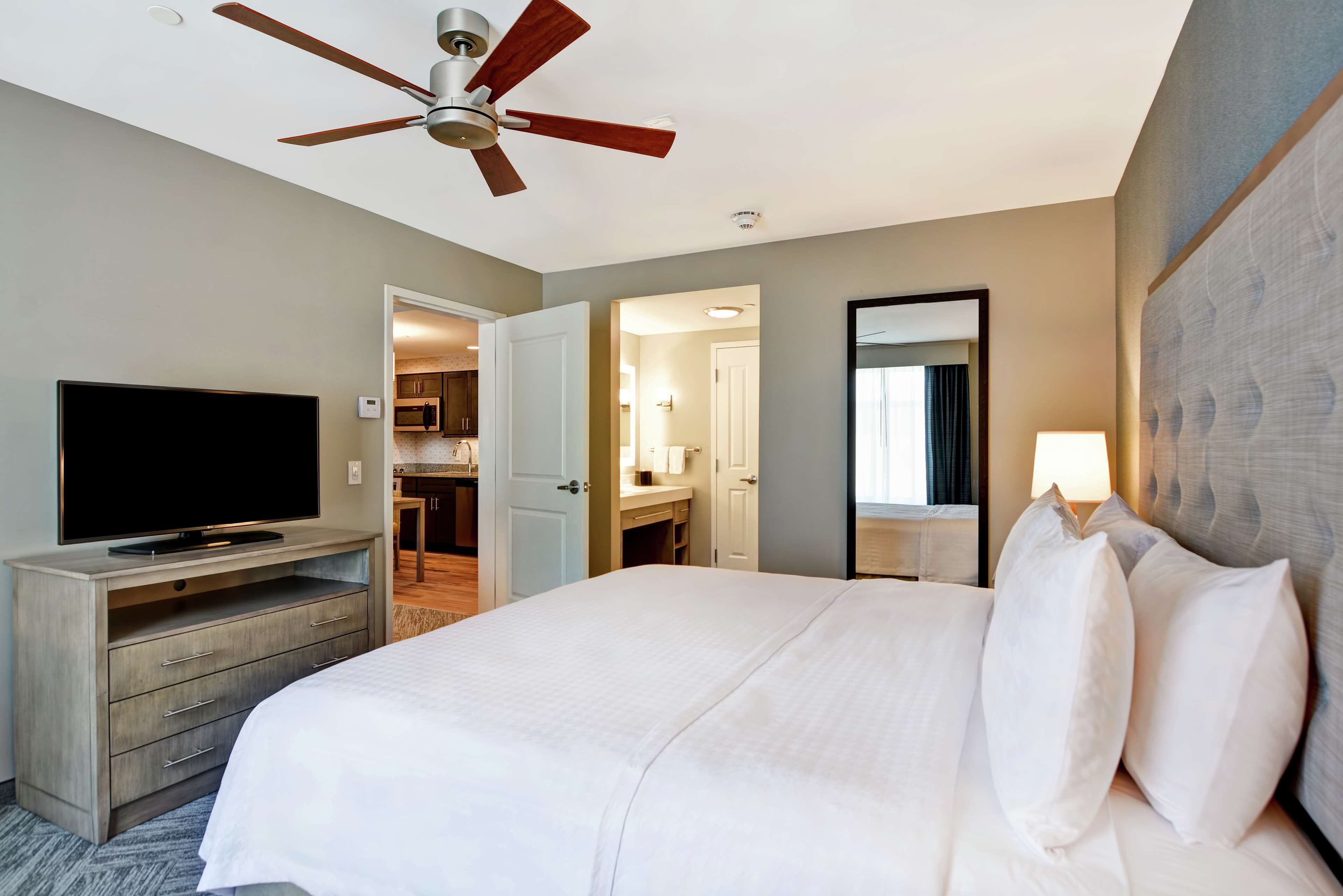 Habitación de hotel con cama, televisor en una cómoda de madera, ventilador de techo y baño visible a través de una puerta abierta.