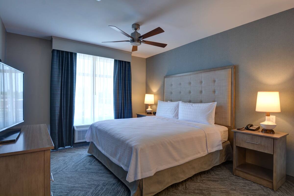A hotel room with a large bed, two bedside tables with lamps, a ceiling fan, a TV on a stand, and a window with curtains.