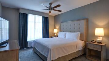 A hotel room with a large bed, two bedside tables with lamps, a ceiling fan, a TV on a stand, and a window with curtains.