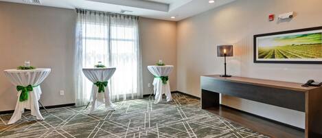 A hotel room with a geometric patterned carpet, two small round tables with green sashes, a large picture on the wall, and a console table with a lamp.