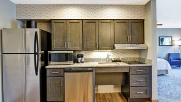 A compact kitchen with a stainless steel refrigerator, a built-in microwave, and a dishwasher. There is a granite countertop, dark wooden cabinets, and a small sink.