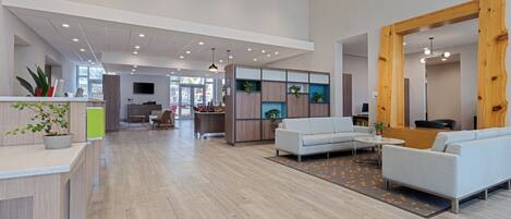 A spacious, modern interior with a wooden reception desk, a seating area with sofas, and a kitchen in the background.