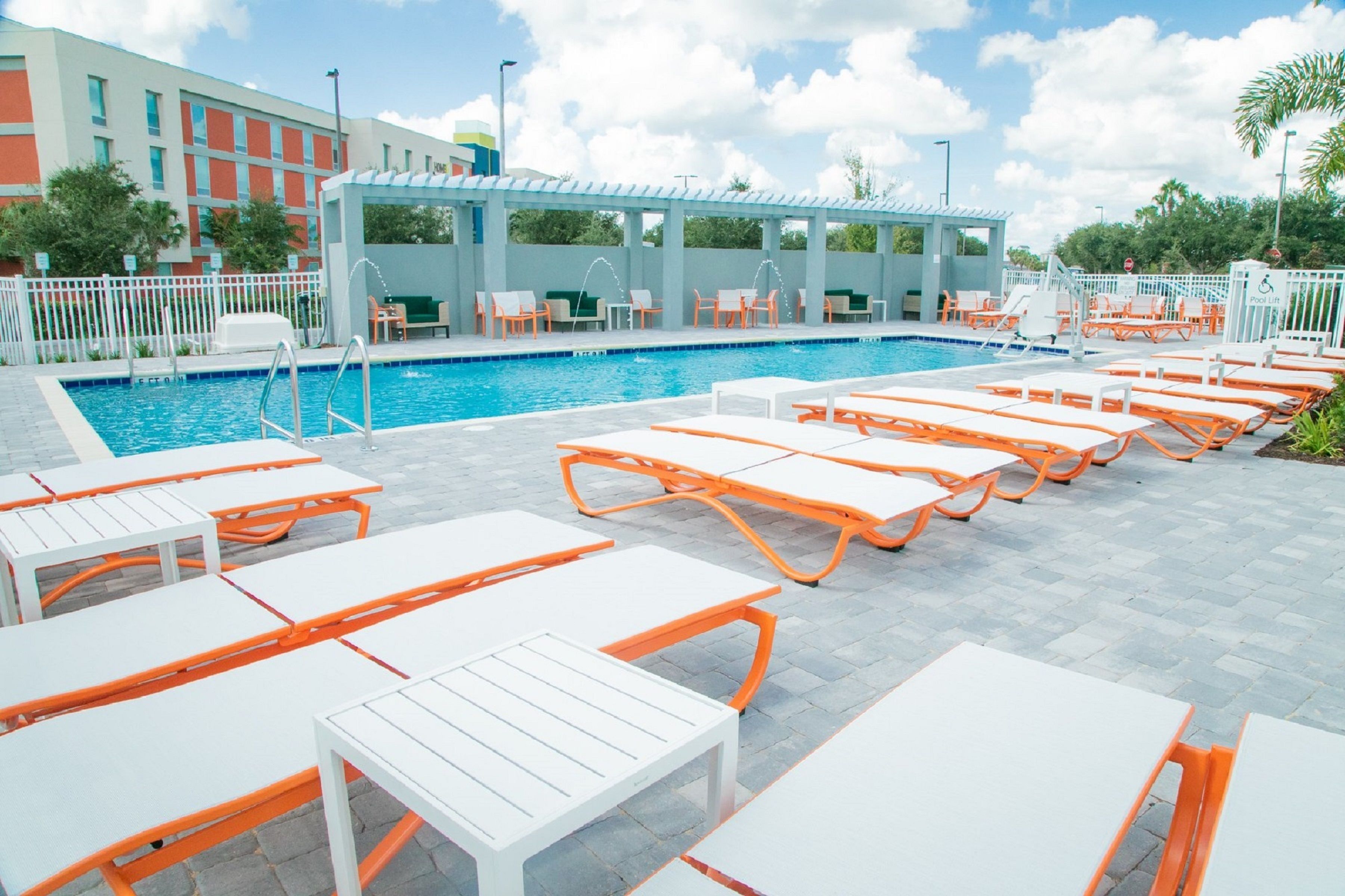 A poolside area with white and orange lounge chairs and tables, a clear blue pool, and a covered seating area with umbrellas.