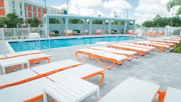 A poolside area with white and orange lounge chairs and tables, a clear blue pool, and a covered seating area with umbrellas.