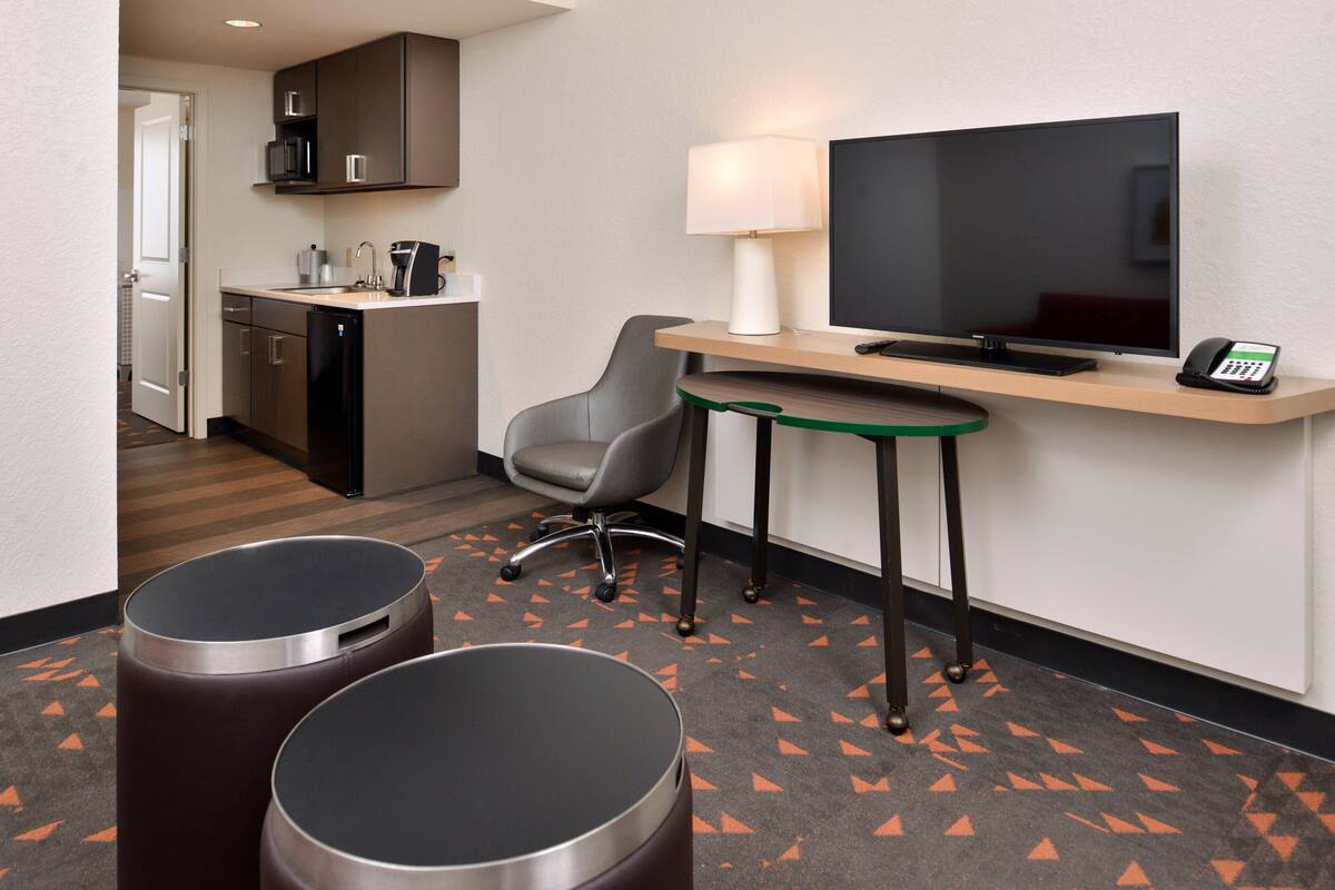 Een hotelkamer met kitchenette, televisie, bureau met lamp, twee afvalbakken en een stoel.