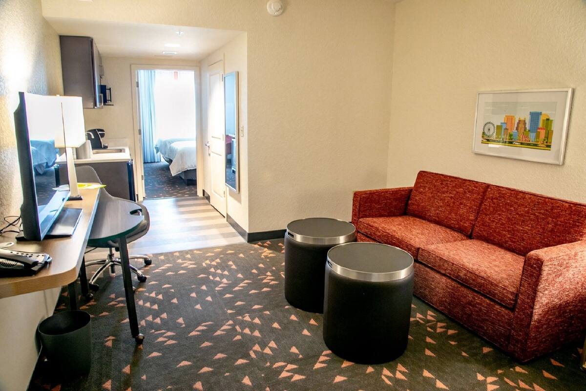 A hotel room with a desk, computer, two trash bins, a red sofa, and a cityscape picture on the wall.