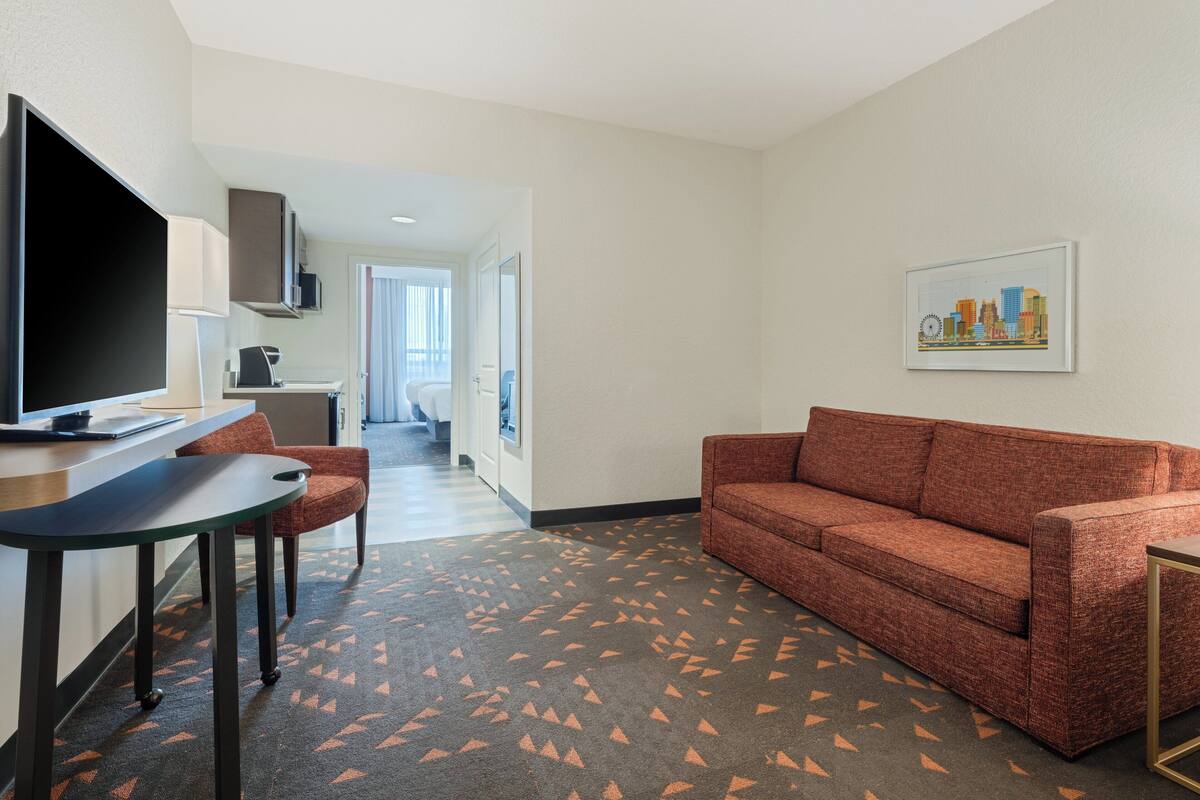 A hotel room with a flat-screen TV, a sofa, a desk with a lamp, and a patterned carpet.