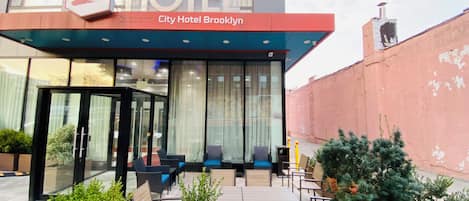 The exterior of City Hotel Brooklyn with a modern design, featuring a prominent 'L' logo, outdoor seating area with tables and chairs, and potted plants.