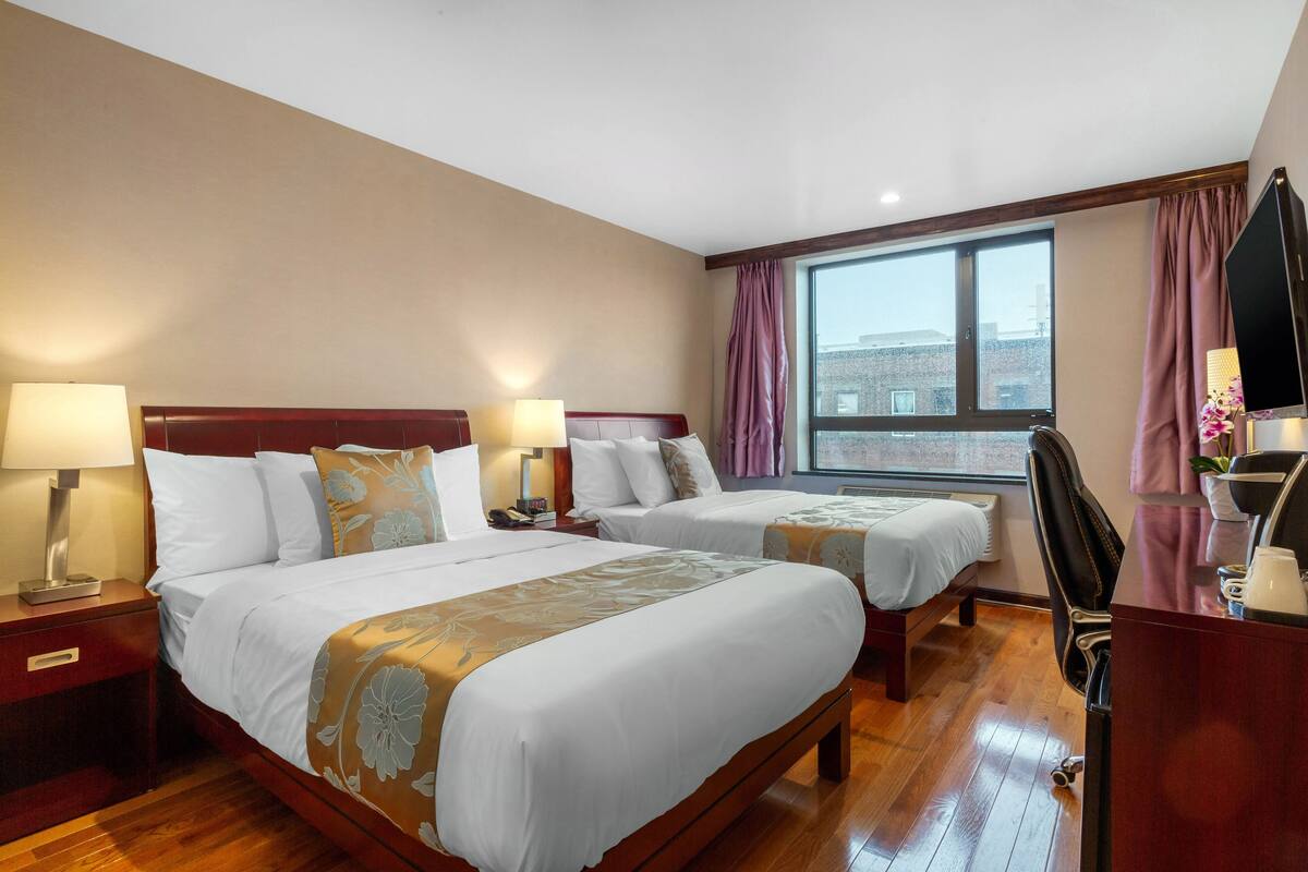 A hotel room with two beds, wooden flooring, a desk with a chair, a window with curtains, and a TV.