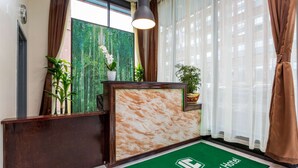 Zona de recepción con un mostrador de madera, un felpudo con el nombre del hotel, un ventanal con vistas a edificios y una lámpara colgante.