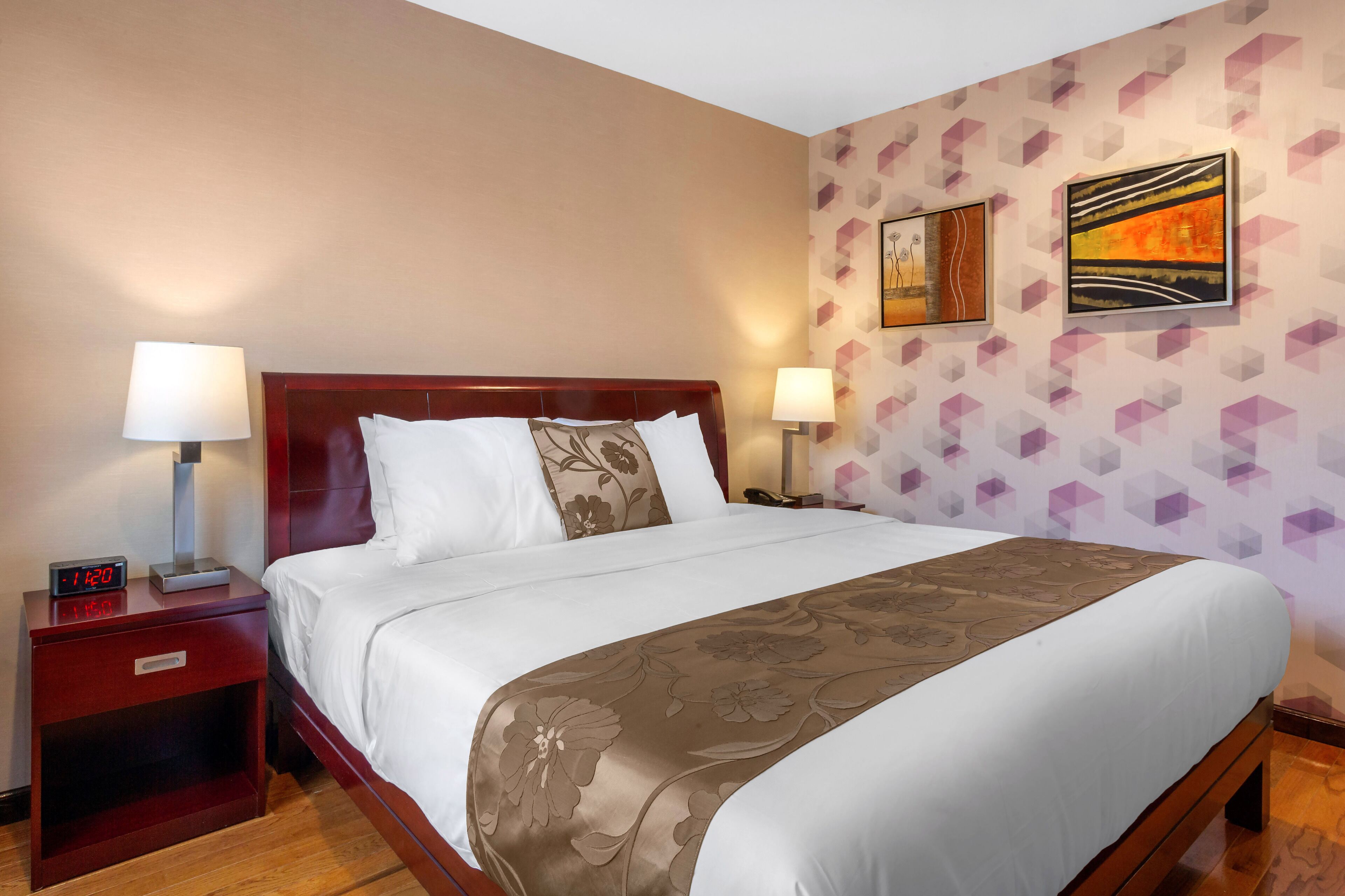 A hotel room with a large bed, two bedside tables, a nightstand, and a wall with geometric-patterned wallpaper and two paintings.